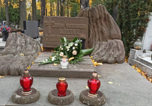 Zdjęcie samego grobu: widoczna tablica, wiązanka kwiatów i biały znicz na podwyższeniu oraz trzy czerwone znicze trochę niżej.