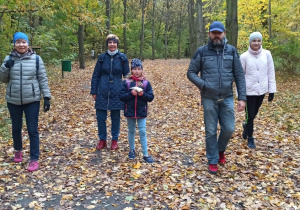Dwie panie, jeden pan i dwie dziewczynki idą alejką parkową, pod ich nogami gruba warstwa kolorowych lisci.