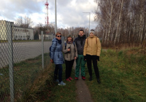 Przy ogrodzeniu z siatki stoją cztery uśmiechnięte postacie. Po prawej stronie znajduje się lasek brzozowy, po lewej białe zabudowania.