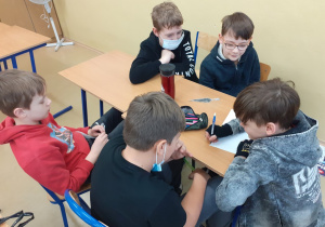 Dzieci siedzą przy ławce i pracują w grupie , zapisują na kartce wymyśloną przez siebie historyjkę.