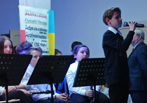 Chłopiec stojący na scenie i recytujący wiersz przez mikrofon. W tle orkiestra szkolna.