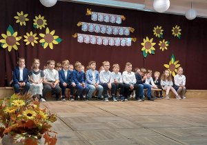 Uczniowie klasy IIa ubrani na galowo siedzą na scenie przy dekoracji z okazji Święta Edukacji Narodowej.