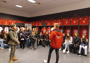 Szatnia Reprezentacji Polski w piłce nożnej, młodzież siedząc lub stojąc z uwagą słucha ciekawostek o tym miejscu przekazywanych przez pana przewodnika.