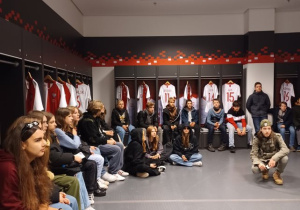 Szatnia Reprezentacji Polski w piłce nożnej, młodzież siedząc lub stojąca z uwagą słucha ciekawostek o tym miejscu przekazywanych przez pana przewodnika.