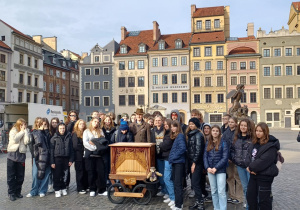Zdjęcie grupowe wykonane z panem kataryniarzem, który z katarynką znajduje się w centralnej części, dookoła roześmiani uczniowie.