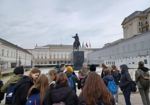 Młodzież słuchająca ciekawostek opowiadanych przez panią przewodnik stojącą przed Pałacem Prezydenckim.
