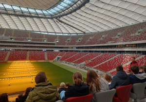 Młodzież siedząca na trybunach Stadionu Narodowego słuchająca pana przewodnika.