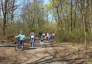 Parkowa aleja. Po lewej stronie zdjęcia stoi drewniana ławeczka. Obok niej przechodzi pani Małgosia prowadząc rower. Tuż za nią maszeruje zwarta grupa spacerowiczów. W tle, jeszcze bezlistne drzewa.
