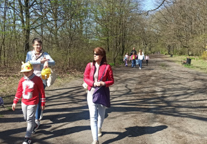 Po ścieżce parkowej maszerują dwie grupy spacerowiczów. Na czele pierwszej idzie dwóch chłopców, a tuż za nimi panie w okularach. Na ziemi ścielą się cienie ludzi i drzew.