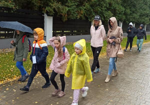 Mokra ulica wyłożona kostką brukową, która błyszczy się od deszczu. Na pierwszym planie czwórka dzieci – dwie dziewczynki i dwóch chłopców, wszyscy mają na głowach kaptury. Chłopiec po lewej stronie dodatkowo niesie nad sobą rozłożony granatowy parasol. Za nimi dwie panie. Jedna w kurtce z kapturem, a druga ma na głowie czapkę bejsbolówkę z napisem ICON. W oddali widać dwóch chłopców. Obaj mają granatowe kurtki z kapturami, jeden z nich ma dodatkowo czapkę z daszkiem wystającą spod kaptura. Wszyscy podążają wzdłuż trawnika, za którym jest ogrodzenie posesji.