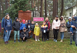 Zdjęcie grupowe. Osiemnaście osób pozuje do zdjęcia na trawniku przed ogrodzeniem, na którym widać sporych rozmiarów tabliczkę z napisem MOLLA. I tak od lewej stoją: kobieta w bordowej kurtce i seledynowych spodniach, obok młoda kobieta w granatowej kurtce i dżinsach, za nią, nie całkiem widoczny, wysoki mężczyzna również w granatowej kurtce i dżinsach. Dalej stoi ośmioro dzieci – sześciu chłopców i jedna dziewczynka, większość z nich ma osłonięte kapturami głowy. Po środku grupy – tuż pod napisem stoi dziewczynka w żółtym płaszczyku przeciwdeszczowym. Po prawej stronie tabliczki widać panią z chłopcem, który ma na szyi zawieszkę. Dalej po prawo stoją dwie panie – jedna w czapce, a druga w kapturze, obok nich pan w dzianinowej czapce trzymający na rękach chłopca, a za nimi widać czubek głowy mężczyzny w okularach.