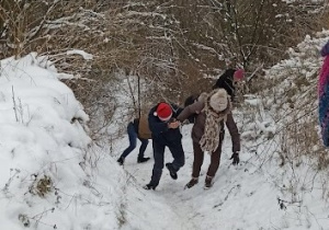 Kilka osób wspinających się stromą ośnieżoną ścieżką. Na pierwszym planie kobieta w szarej czapce z pomponem i trzymający ja za rękę mężczyzna w czerwonej czapce Mikołaja.