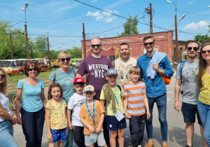 Pięcioro dzieci w letnich strojach pozuje do zdjęcia przed bramą Zajezdni Muzealnej BRUS. W dali, za bramą widać zabudowania z czerwonej cegły.
