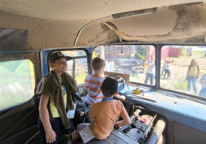 Trzech chłopców, z których dwóch ma na głowie czapki z daszkiem, siedzi w kabinie autobusu muzealnego typu "ogórek", chłopiec bez czapki siedzi tuż przy kierownicy. Za szybą, przed autobusem widać czworo dorosłych.
