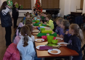 Dzieci siedzą przy długim stole, na którym znajdują się artykuły papiernicze i ozdoby. Prowadzące zajęcia prezentują przykładowe prace – wieńce adwentowe.