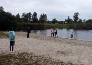 Cztery dziewczynki i troje dorosłych na piasku nad stawem, po którym pływają kaczki.