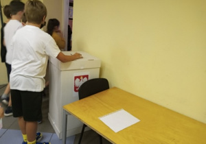 W sali lekcyjnej uczeń wrzuca swój głos do urny wyborczej.