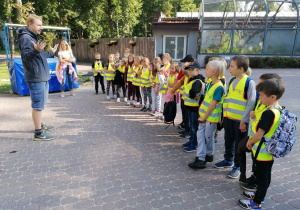 Uczniowie klasy IIc ubrani w kamizelki odblaskowe, stoją naprzeciw przewodnika, który przedstawia im plan i trasę wspólnego zwiedzania ogrodu zoologicznego.