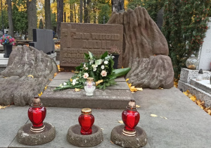 Zdjęcie samego grobu: widoczna tablica, wiązanka kwiatów i biały znicz na podwyższeniu oraz trzy czerwone znicze trochę niżej.