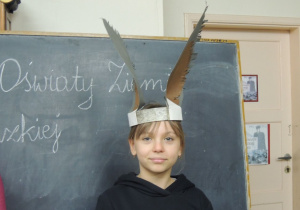 Na tle czarnej tablicy szkolnej stoi uczennica z przepaską na głowie imitującą ośle uszy.