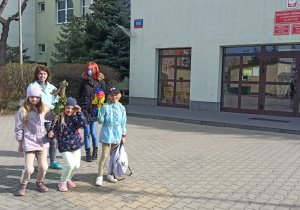 Cztery dziewczynki i jedna nauczycielka stoją przed budynkiem szkoły. Dwie z nich trzymają w rękach kukiełki marzanny.