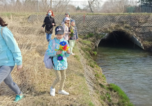 Cztery dziewczynki i dwie kobiety idą wzdłuż brzegu niewielkiej rzeki. Dwie dziewczynki trzymają w rękach kukiełki.