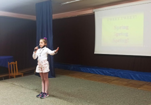 Dziewczynka stoi na scenie trzymając w dłoni mikrofon recytuje wierszyk, drugą ręką gestykuluje.