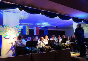 Zdjęcie przedstawiające orkiestrę na scenie. Na pierwszym planie dziewczęta grające na fletach oraz Pan Dyrektor (dyrygent). W tle falaga Ukrainy, Polski, Unii Europejskiej.