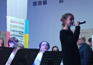 Dziewczynka stojąca na scenie i recytująca wiersz przez mikrofon. W tle orkiestra szkolna.