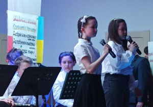 Dziewczynki stojące na scenie i recytujące wiersz przez mikrofon. W tle orkiestra szkolna.