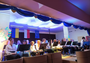 Zdjęcie przedstawiające orkiestrę na scenie. Po prawej stronie na krzesełku siedzi chłopiec grający na gitarze.W tle falaga Ukrainy, Polski, Unii Europejskiej.