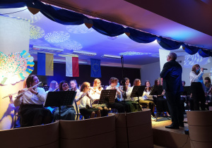 Zdjęcie przedstawiające orkiestrę na scenie. Na pierwszym planie dziewczęta grające na fletach, Pan Dyrektor (dyrygent) oraz Dziewczynka śpiewająca piosenkę przez mikrofon. W tle falaga Ukrainy, Polski, Unii Europejskiej.