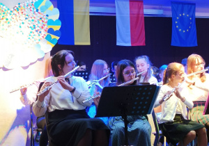 Zdjęcie przedstawiające orkiestrę na scenie. Na pierwszym planie dziewczęta grające na fletach. W tle falaga Ukrainy, Polski, Unii Europejskiej.