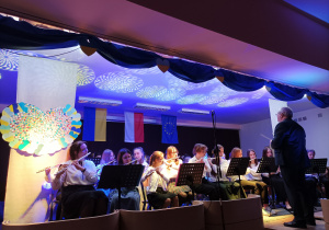 Zdjęcie przedstawiające orkiestrę na scenie. Na pierwszym planie dziewczęta grające na fletach oraz Pan Dyrektor (dyrygent). W tle falaga Ukrainy, Polski, Unii Europejskiej.