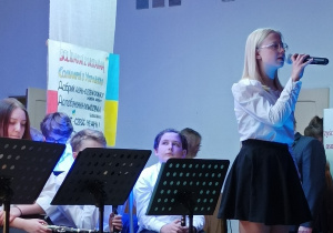 Dziewczynka stojąca na scenie i recytująca wiersz przez mikrofon. W tle orkiestra szkolna.