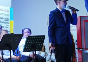Chłopiec stojący na scenie i recytujący wiersz przez mikrofon. W tle orkiestra szkolna.