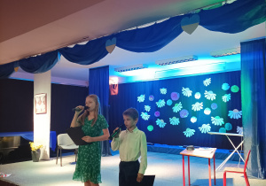 Chłopiec i dziewczynka stojący na scenie i przemawiający do gości przez mikrofon.