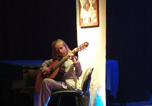 Helena Jóźwicka siedząca na krześle na scenie i grająca na gitarze.