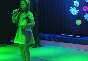 Dziewczynka stojąca na scenie, recytująca wiersz. W jednej rece trzyma pluszowego misia, w drugiej mikrofon.