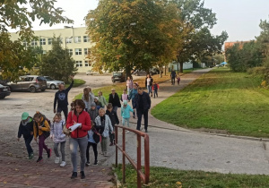 Grupa dzieci i dorosłych wyrusza na wycieczkę, w tle budynek szkoły, na przedzie nauczycielka w czerwonej kurtce.