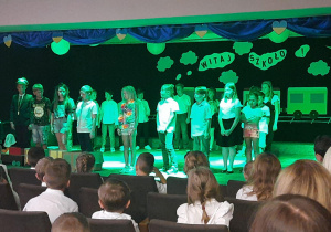Na scenie grupa dzieci w wakacyjnych i szkolnych strojach. uczeń stojący pośrodku w pierwszym rzędzie mówi do mikrofonu. na pierwszym planie widzoawie.