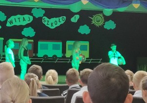 Na scenie stoją trzy dziewczynki i chłopiec, w strojach wakacyjnych. dziewczynki zwrócone są w stronę chłopca, który trzyma piłkę i mówi do mikrofonu. W tle dekoracja z pociagiem, chmurkami i słońcem.