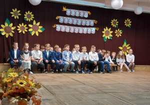 Uczniowie klasy IIa ubrani na galowo siedzą na scenie przy dekoracji z okazji Święta Edukacji Narodowej.