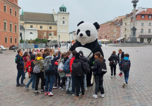 Stare Miasto w Warszawie. Uczniowie stoją wokół dużego misia pandy, w tle Kolumna Zygmunta III Wazy.