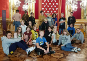 Zamek Królewski w Warszawie – Sala Tronowa. Dzieci siedzą na podłodze i słuchają przewodnika. Za dziećmi tron oraz lustra.