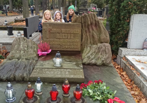 Za płytą pomnika z napisem Jan Witold Moll profesor medycyny i kardiologi stoją trzy uśmiechnięte dziewczynki. Po prawej stronie nagrobka leży bukiet czerwonych róż. Z przodu stoją zapalone biało-czerwone znicze.