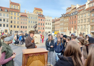 Uczniowie stojący na Rynku Starego Miasta wpatruje się w pana katarynarza, który tłumaczy jak działa mechanizm katarynki.