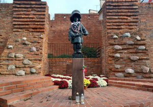 Pomnik Małego Powstańca, przed nim znajdują się znicze, za nim donice wypełnione kwiatami w barwach narodowych.