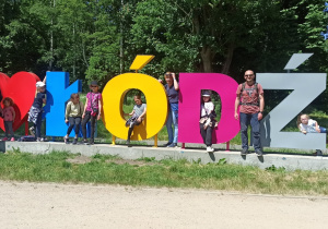 Siedem dziewczynek, jedna kobieta i dwóch mężczyzn pozuje do zdjęcia na tle napisu "Ja kocham Łódź". Słowo "kocham" przedstawia czerwone serce.