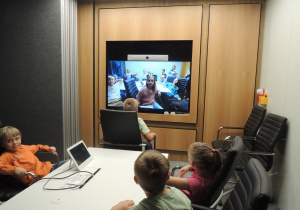 Grupka dzieci siedzi w sali konferencyjnej przed włączonym dużym ekranem ściennym, na którym widoczne są dzieci z drugiej grupy przebywające w innym pomieszczeniu.